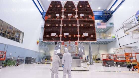 Three large solar panels hang in the back of a cleanroom warehouse room where two workers dressed in white suits stand in the foreground — Image credit: The Nancy Grace Roman Space Telescope, NASA's next great observatory, is finally complete / https://www.space.com/author/monisha-ravisetti