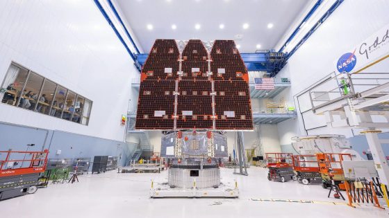 inside a white hangar with a large ceiling sits a metal cylinder from which extend three long rectangular solar panels, all pointing towards the ceiling — Image credit: NASA reveals its Roman Space Telescope today: How to watch live, and what's next for the next-generation observatory / https://www.space.com/author/elizabeth-howell