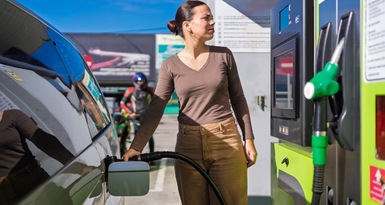Woman refuels her car at a gas station on a sunny day, pondering rising gas prices. — Image credit: Who Controls Gas Prices in the US? / https://www.kiplinger.com/author/jim-patterson
